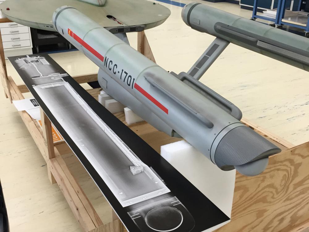 The Starship Enterprise, a gray-colored disc-shaped starship studio model with two rear, cylinder-shaped sections, is placed above an X-ray scan print of one of the rear sections of the model.