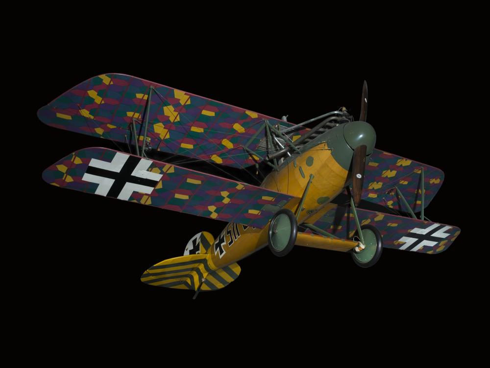 Biplane with multi-color, camouflaged wings, wood grain finish fuselage, and green and yellow details on tail and nose, seen from below