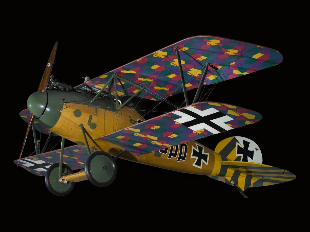 Side view of biplane with multi-color, camouflaged wings, wood grain finish fuselage, and green and yellow details on tail and nose