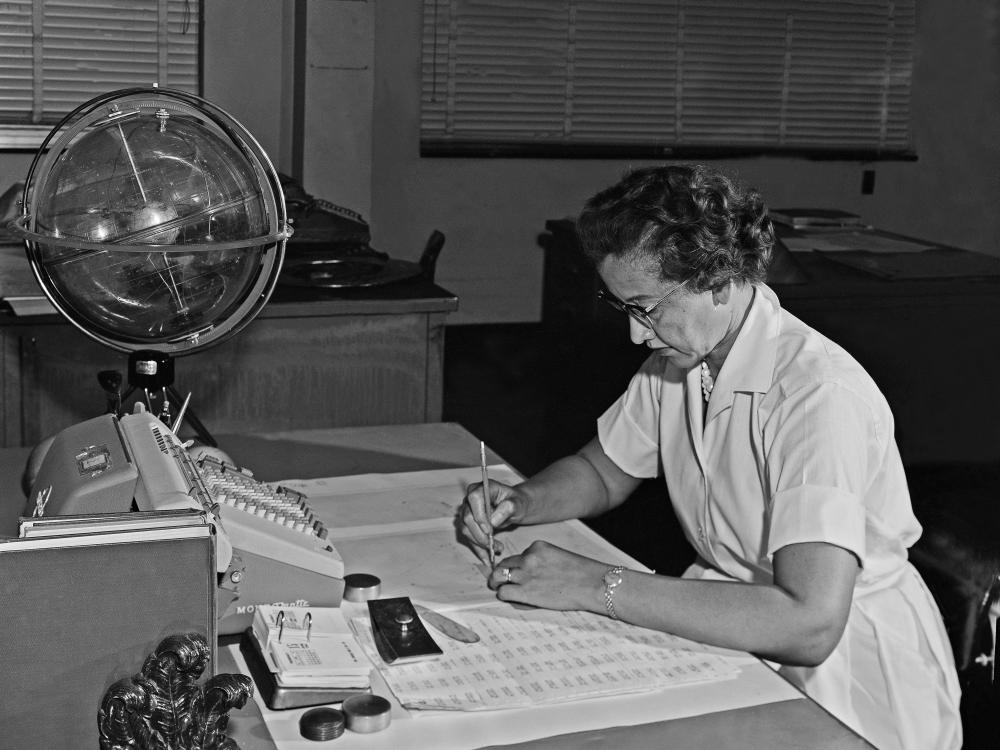 A woman in a collared shirt and necklace leans over her desk working.