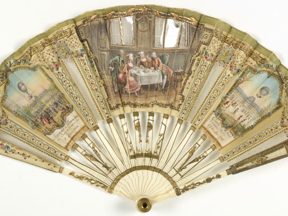 Ornate, gilded ivory decorative fan with depictions of balloon launches on the left and right sections and a center section of people viewing a balloon through a window.