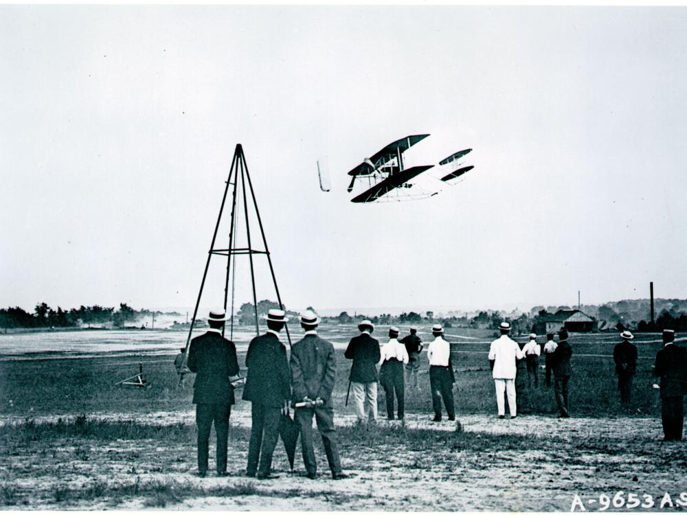 Black and white photo of the Wright Flyer in flight with crowd of onlookers below. 