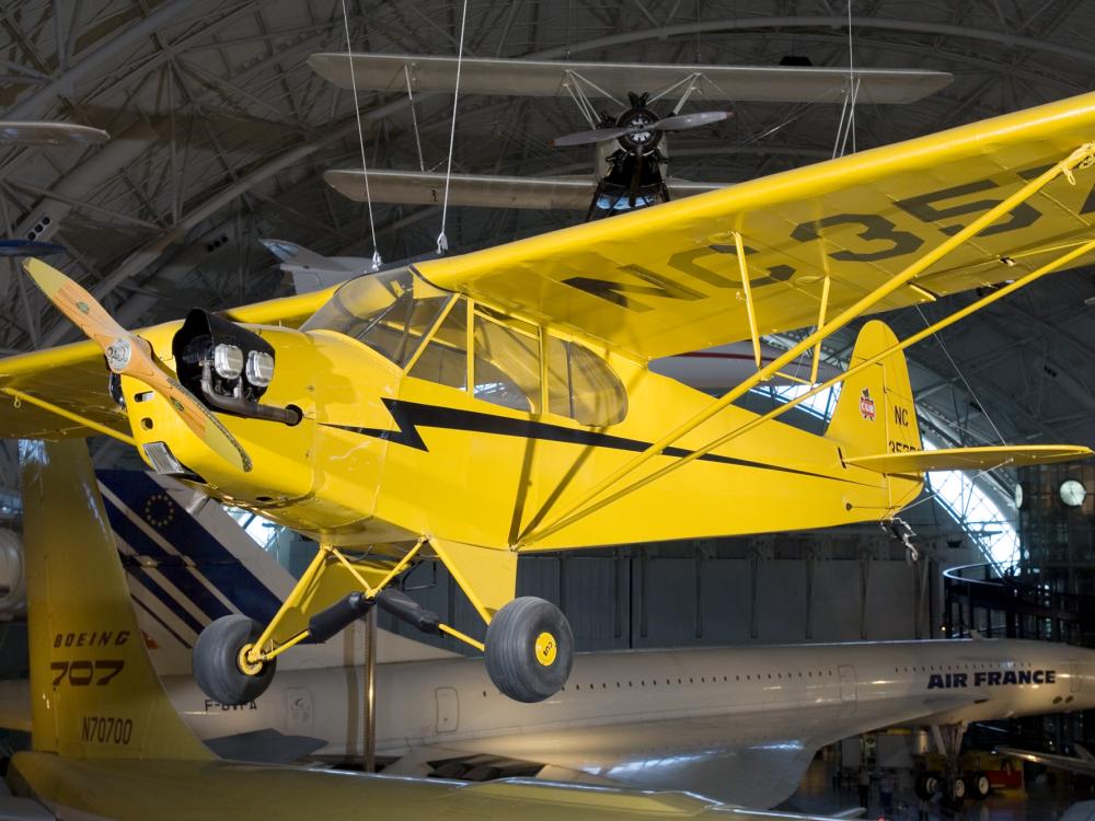 A Piper J-3, a yellow monoplane with one engine, is displayed from a suspended position inside the Museum.