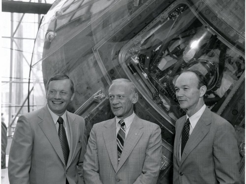 Black and white of Armstrong, Aldrin, and Collins stand beside the Apollo 11 command module.