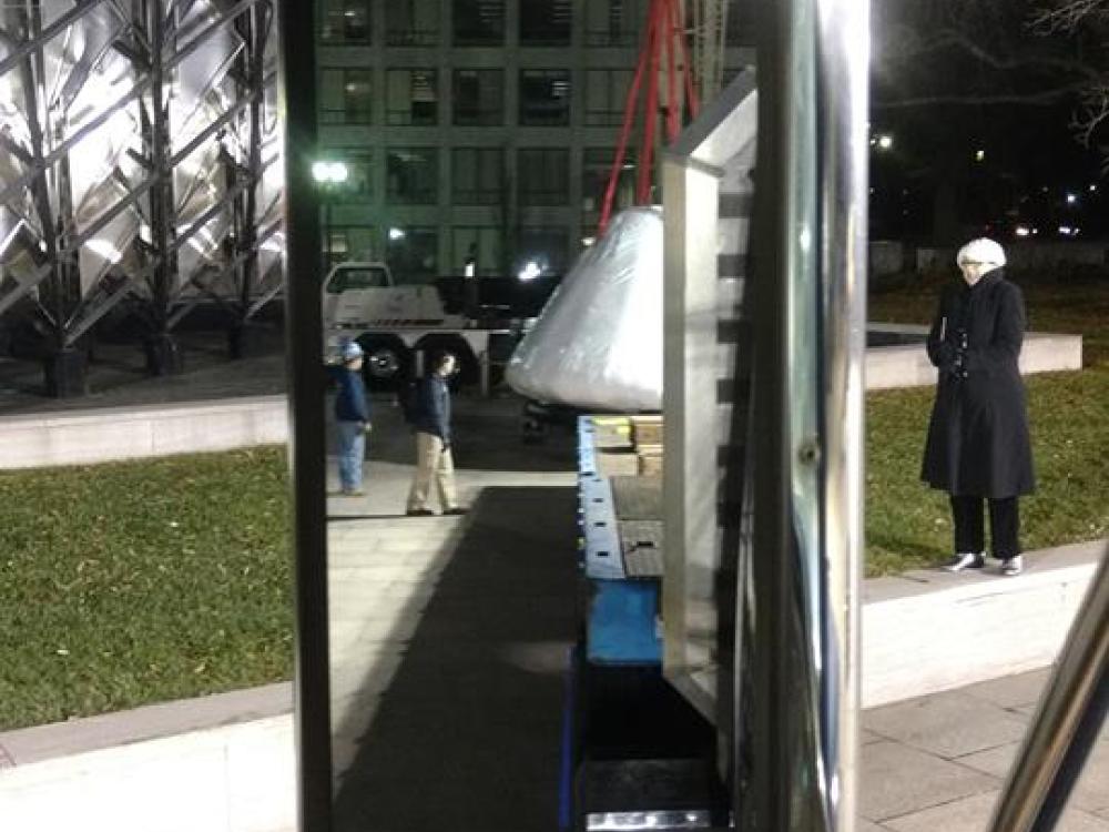 Image of a rear view mirror. In the mirror you can see the outline of the command module on a truck flat bed. 