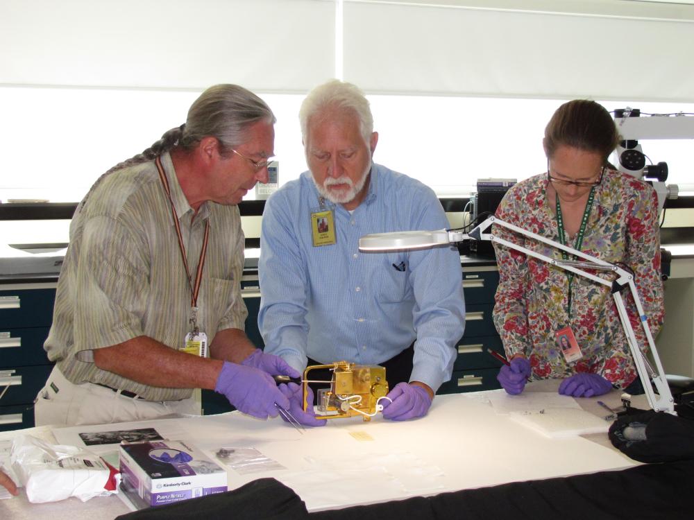 Two men work together on taking a part a film transport located on a bench. Beside them is a woman who tracks each componenet. 