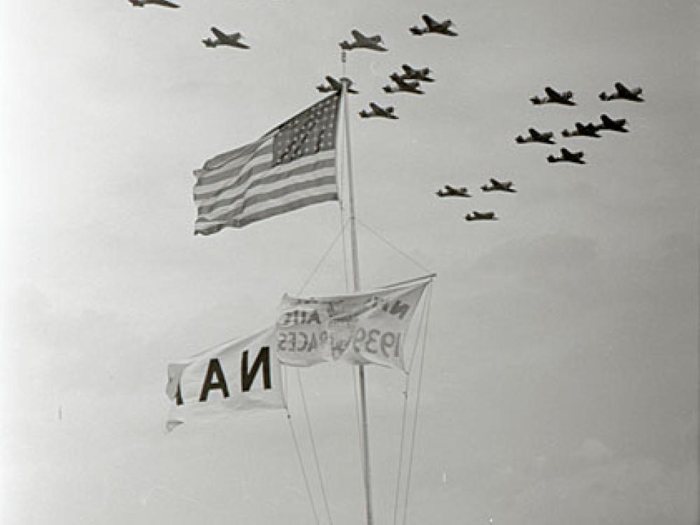 Formation of airplanes fly past American flag. Spectators can seen at bottom.