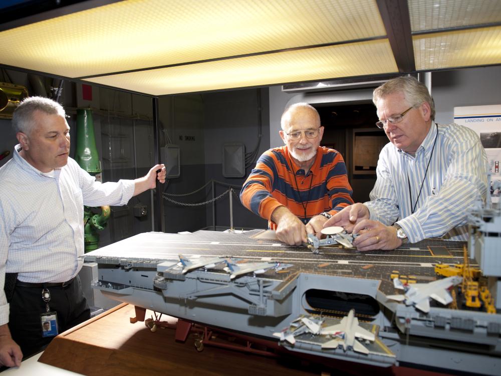 curator Chris Moore (left) looks on as Steve Henninger (center), assisted by Paul Moore, frees the E-2C on the model of the USS Enterprise.