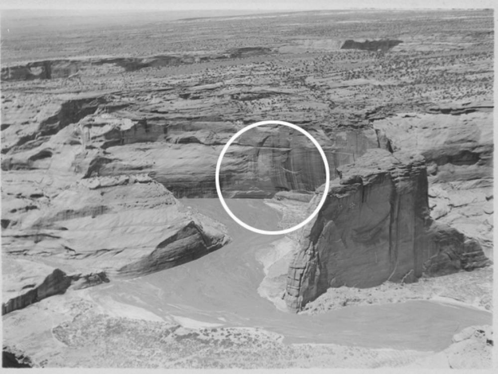 A black and white photograph showing large vertical rock formations that house cliff dwellings. The dwelling area is circled by a superimposed white circle.