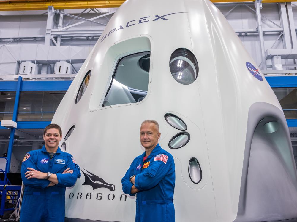 two astronauts in blue flight suits in from of capsule