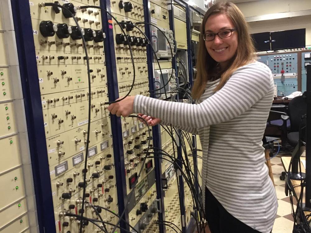 Woman stands in front of a large control board with multiple wires and access points. 