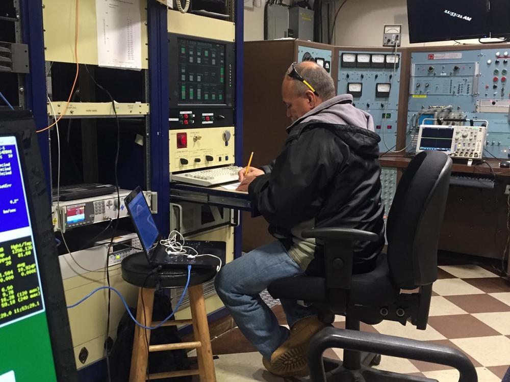 Man works at a small desk surrounded by equipment. 