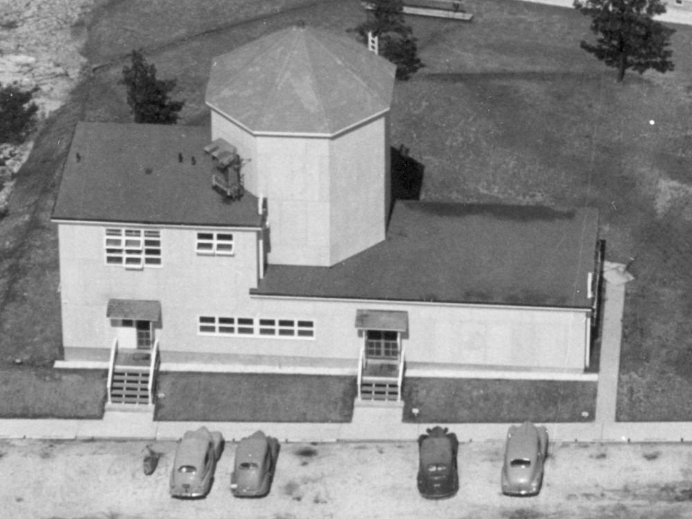 Aerial photo of a building with one large silo and cars parked out front. 