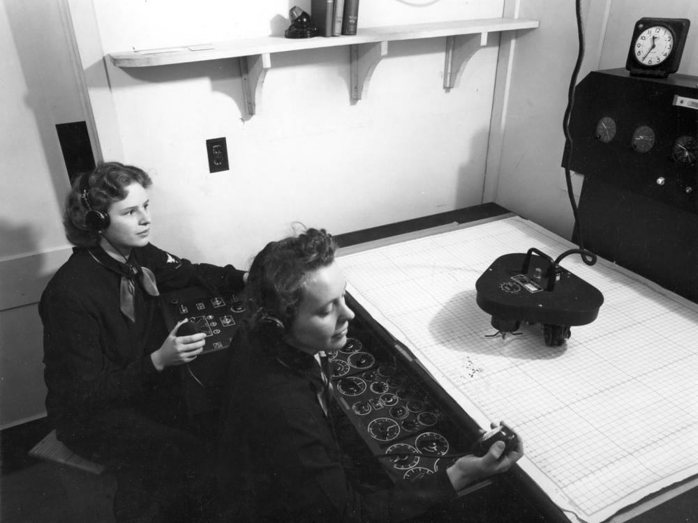 Two women sit in front of controls and plotting table. 