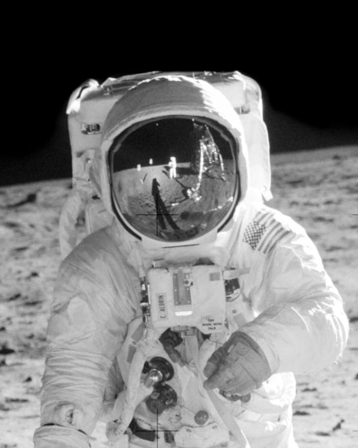 A photograph of a man in a spacesuit on the moon. IN the reflection of the helmet you can see another astronaut and the spacecraft.