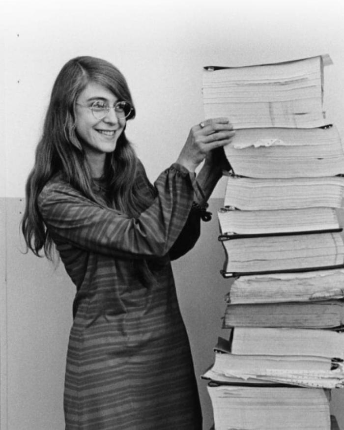 A woman stands next to a stack of books as tall as she is.