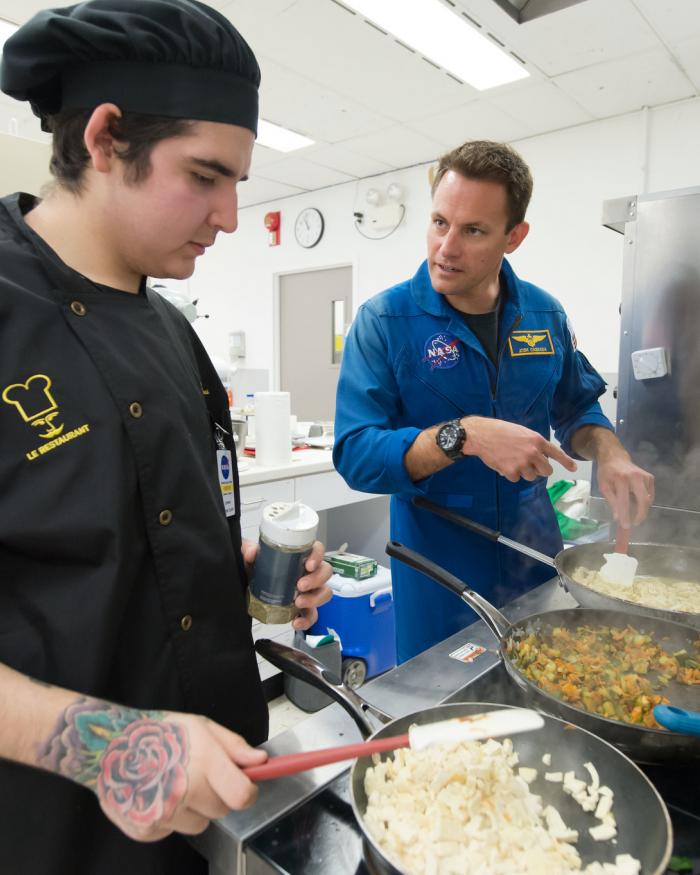 Chef in black chef coat cooks while an astronaut in blue flight suit helps