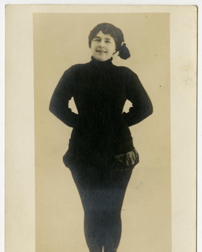 A woman in a jaunty hat stands with her hands behind her back. 