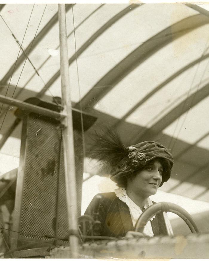 A woman in a hat with three pins and a feather behind a steering wheel.