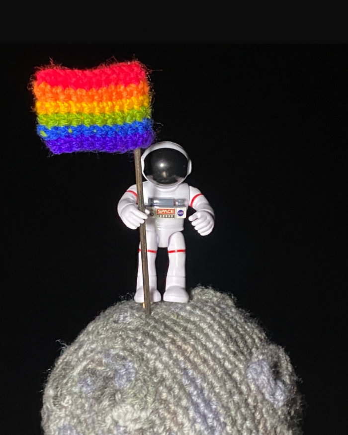 Astronaut standing on crocheted Moon holding a Pride flag