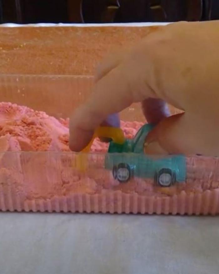 A hand moves a toy truck through pink kinetic sand in a plastic container. 