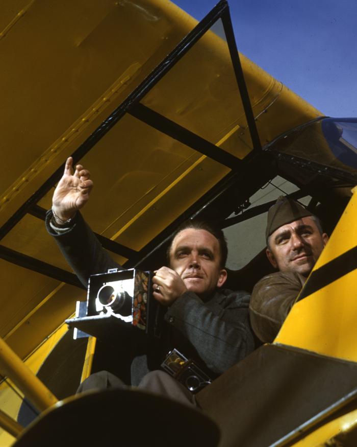 A photo taken from underneath an aircraft. One man leans out with a camera, the other is a pilot.