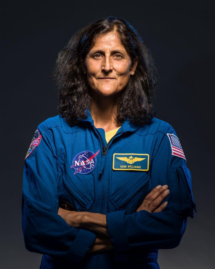 NASA Astronaut Sunita Williams looks at the camera, with a dark background, in a blue suit. 