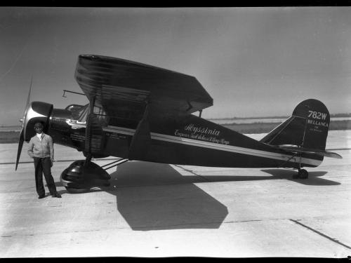 Black and white left side view of monoplane painted black with white stripe down middle. Cursive handwritten text in center of plane reads: "Abyssinia Emperor Haile Salassi I King of Kings." Tail text "782W/Bellanca" Black man by nose right hand in pocket