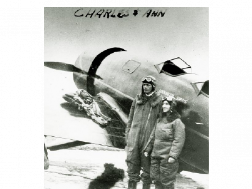 Charles and Anne Lindbergh stand on side of their Lockheed Sirius while wearing aviator gear.