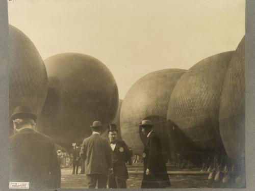 Black and white photo. In the center are three men dressed in suits, the two in the right wearing derbys, the middle man in a top hat. Five hot air balloons in the background. The back of a man in a suit and derby is in the foreground in the lower left corner.