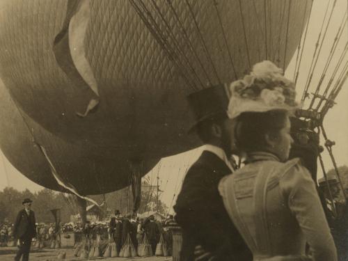 A sepia tone photograph, in the foreground are fashionably dressed people from the early 1900s, in the background are hot air balloons. 