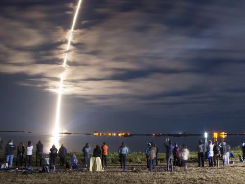 Spectators gathered by a body of water and watching a rocket launching to the sky leaving behind a glowing streak as it enters the clouds.