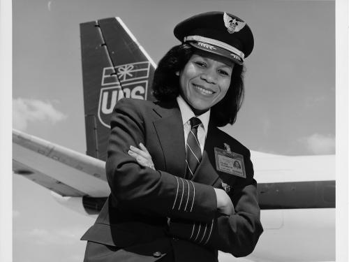 Black & white photograph. African-American woman with hair to shoulders turned to her right with left hand crossed under right.  She is wearing a dark uniform with three pale thin stripes above each hand and an ID badge on her left chest.  She has a white collared shirt with a dark tie with thin pale stripes under the hack. She wears a stiff pilots cap with two laurel leaves on the brim and a star surrounded by wings on the crest.  In the background left is an airplane tail with the UPS logo of wrapped pack