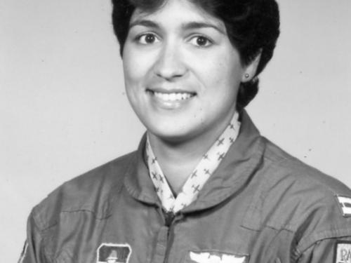 Black and white portrait of woman with short wavy hair, parted in the middle.  Stud earring in left ear (right ear is not visible).  She is wearing a dark jacket with a zipper with a cloth pull.  Patch on her right side (the left of the image) features a icon with wings.  Two patches on her left side (right of photo). Top is wings. Bottom is a nameplate that says "Custodio."  There are two pens in her left sleeve. Another patch is cut off by the edge. White collar with a dark airplane print under the jacket