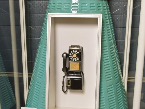 A phonebooth in turquoise color that is shaped like a space capsule with a dial phone in the middle.