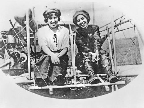 Two sisters sit in an airplane, laughing and smiling.