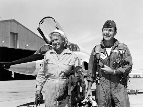 Black and white image of Jackie Cochran and Chuck Yeager walking and smiling with an aircraft in the background.