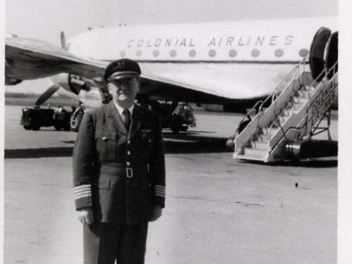 Black and white photograph. Foreground: Man in dark pilot's uniform with a cap and stripes at the ends of his sleaves. Background: Side of a passenger airplane labelled "Colonial Airlines" above circular windows. A set of airstairs is behind and to the right of the man.