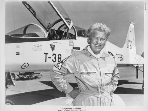 An aged Jackie Cochran posing with her hands on her hips and smiling and a T-38 Talon aircraft in the background.