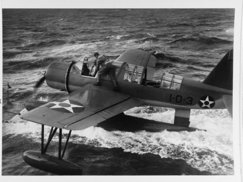 Black and white image of an airplane riding on top of water with a man on its wing.