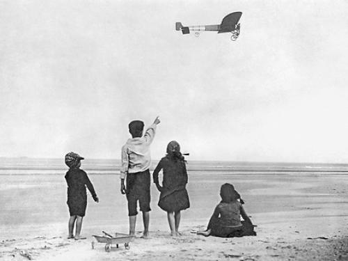 Black and white photo of four children on a beach. One points to an airplane in the distance.