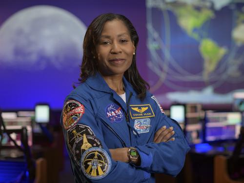 Stephanie Wilson stands with her arms folded, wearing a blue jacket with many space mission patches.