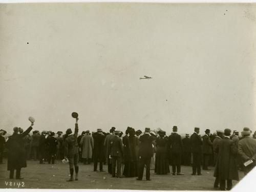 A crowd of people, some cheering, looking toward the sky where an airplane can be seen in the distance.