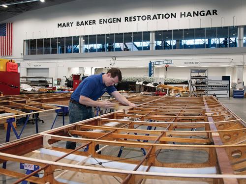 Inside an hangar, a craftsman sands an archival aircraft wing
