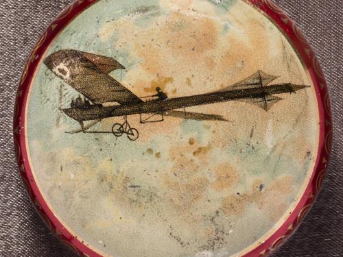 Circular metal box with monoplane and pilot in a cloudy sky painted on the lid. Image surrounded by narrow gold border. Sides and bottom metallic red.