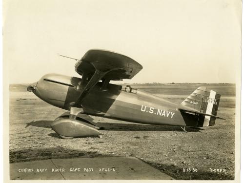 Sepia-toned photo of the left side view of a person in a Curtiss racer monoplane on the ground. The aircraft has U.S. Navy written on the side. 