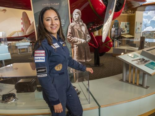 A woman in a flight uniform poses next to a bright colored early aircraft in a museum setting.