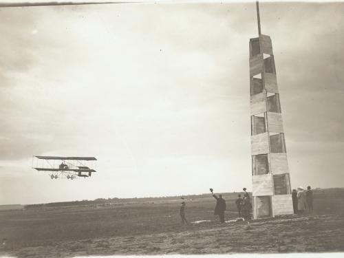 Farman-type biplane rounds a pylon in a field as a handful of people look up, some waving their hats in the air.