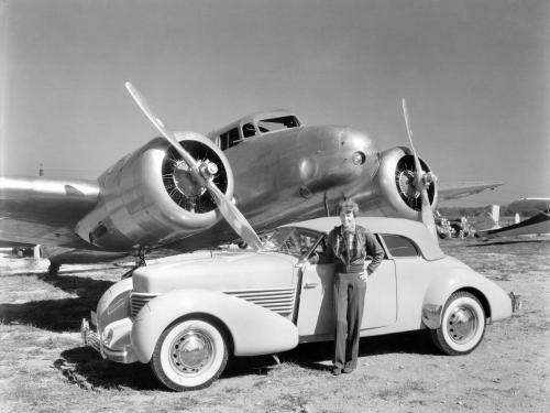 A wideshot of a woman standing next to a car with a aircraft in the background.