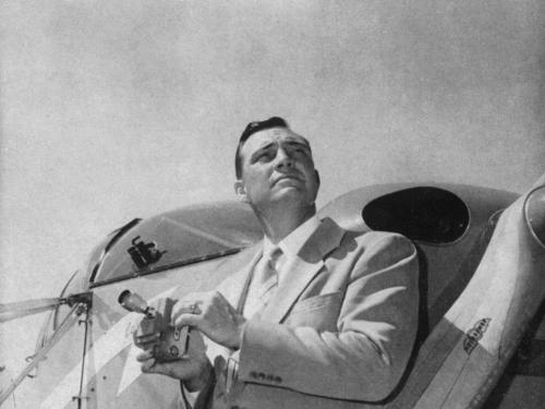 Black and white image of a man standing in front of an airplane, holding an object in his hand, and looking to the right.